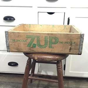 Puede incluir: Caja de madera vintage con la palabra "7UP" en verde, junto con "THE UNCOLA" en los laterales. La caja tiene refuerzos de esquina metálicos y está colocada sobre un taburete de madera marrón. La caja es de madera clara.