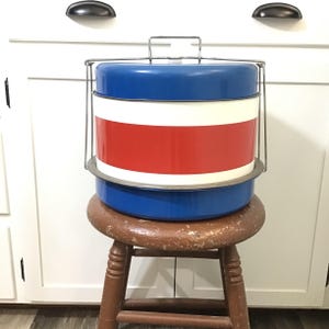 May include: A vintage metal cooler with a blue top and bottom, a white stripe, and a red stripe in the middle. It has a metal handle and is sitting on a wooden stool. The cooler is a classic design.