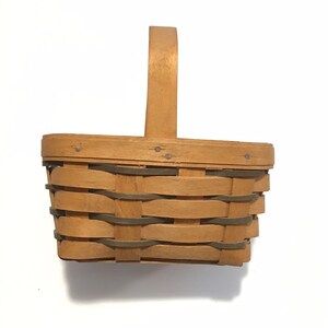 May include: A small, rectangular, woven basket with a wooden handle. The basket is made of natural wood and has a light brown color.