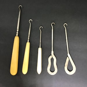 May include: Five antique crochet hooks of varying sizes. The handles are cream-colored, and the hooks are silver-toned. The hooks are arranged on a dark surface, showcasing their different designs and lengths. The hooks are likely used for crafting.