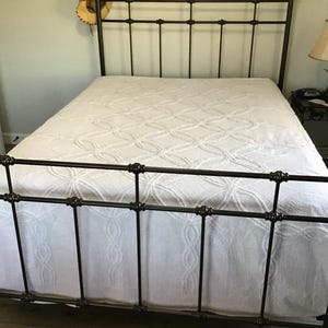 May include: A wrought iron bed frame with a white patterned bedspread and matching bed skirt. A cowboy hat hangs on the headboard. The bed is in a bedroom setting.