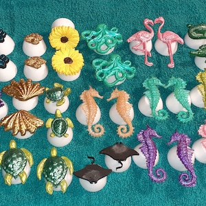 May include: A collection of 28 colorful sea-themed ornaments, including seahorses, turtles, seashells, a starfish, an octopus, a flamingo, a crocodile, and a Mickey Mouse head. The ornaments are displayed on white bases.