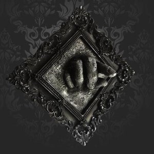 May include: A black ornate frame with a raised, textured, gray hand sculpture inside. The frame has a distressed, antique look.