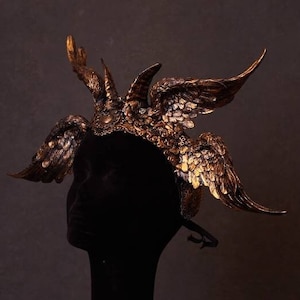 May include: A detailed, gold-colored headpiece with wing-like structures and intricate detailing. The headpiece is displayed on a black mannequin head, set against a dark background. The design evokes a regal or fantasy theme.