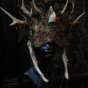 May include: A decorative mask with antlers, a skull, and ornate gold filigree. The mask features a black base with attached antlers, a small skull, and a detailed gold filigree design over the eyes. Bone-like pieces hang from the mask with chain accents.