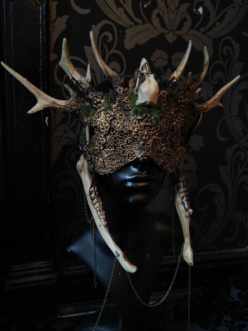 May include: A decorative mask with antlers, a skull, and ornate gold filigree. The mask features a black base with attached antlers, a small skull, and a detailed gold filigree design over the eyes. Bone-like pieces hang from the mask with chain accents.