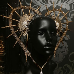 May include: A gold-toned crown with pointed spikes and pearl accents. The crown is worn on a black mannequin head. A gold chain necklace with a pearl pendant hangs from the crown.