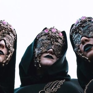 May include: Three individuals wearing black hooded garments and ornate, textured gold-toned masks that cover the upper face. The masks feature intricate filigree details and are accented with small, light pink circular stones. Each person has dark lipstick.