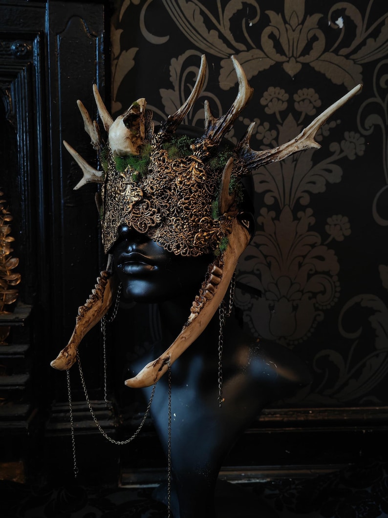 May include: A gothic-style headpiece featuring antlers, a skull, and ornate gold filigree. The crown is adorned with moss and bone elements, with jawbones hanging from chains. The headpiece is displayed on a black mannequin head.