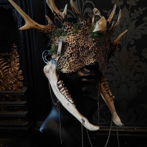 May include: A detailed, ornate headpiece featuring deer antlers, a skull, and a decorative gold-colored mask. The mask is adorned with intricate patterns and is accented with moss. Bone-like elements and chains hang from the sides, creating a gothic aesthetic.
