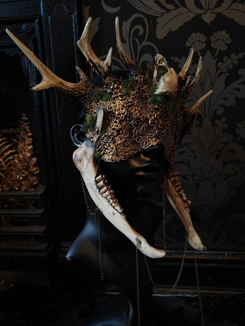 May include: A detailed, ornate headpiece featuring deer antlers, a skull, and a decorative gold-colored mask. The mask is adorned with intricate patterns and is accented with moss. Bone-like elements and chains hang from the sides, creating a gothic aesthetic.