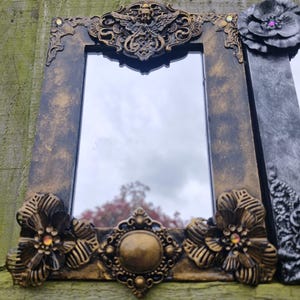 May include: Ornate rectangular mirror with a dark bronze and gold frame. The frame features floral and scrollwork details, with a central decorative element and a large flower on the side. The mirror reflects the sky.