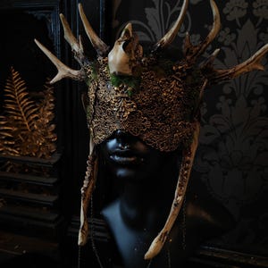 May include: A detailed, ornate headpiece with deer antlers, a skull, and intricate gold filigree. The mask-like design covers the eyes and is attached to a black mannequin head. The headpiece has a dark, gothic aesthetic.