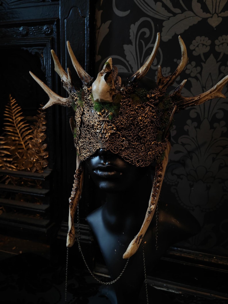 May include: A detailed, ornate headpiece with deer antlers, a skull, and intricate gold filigree. The mask-like design covers the eyes and is attached to a black mannequin head. The headpiece has a dark, gothic aesthetic.