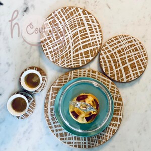 Puede incluir: Posavasos y manteles individuales de corcho con un patrón de cuadrícula blanca. Dos tazas pequeñas de café están sobre posavasos a juego. Un plato azul con un pastel y una tapa de cristal está sobre un mantel individual más grande.