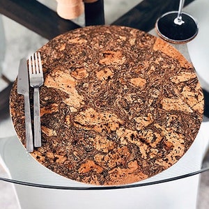 May include: A round cork placemat with a natural, mottled pattern, paired with a fork and knife. The table setting includes small candles and a wine glass. The placemat is approximately 12 inches in diameter.