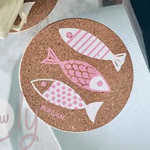 Könnte beinhalten: Korkuntersetzer mit rosa Fischmotiven. Ein Untersetzer hat einen Holzbecher und eine kleine weiße quadratische Schale mit Deckel. Der andere hat drei Fischdesigns und das Wort "PORTUGAL".