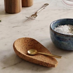 May include: A kitchen scene with wooden oil bottles, a silver fork, a blue ceramic bowl filled with salt, a cork spoon rest with a gold spoon, and fresh herbs on a marble countertop.