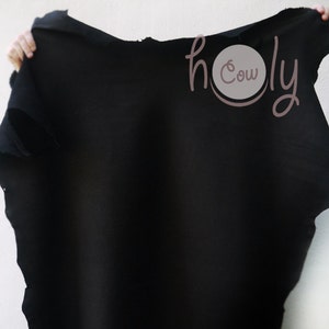 May include: A large, irregularly shaped piece of black leather is held up against a white background. The leather has a rough, unfinished edge. The text "holy cow" is in the upper right corner, in a cursive font.