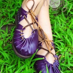 May include: Purple leather sandals with brown laces. The sandals have a cut-out design and are tied at the ankle. The sandals are on a bed of green grass.