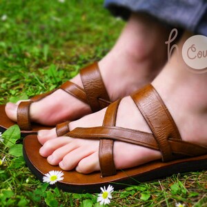 May include: Brown leather sandals with multiple straps crossing over the foot. The sandals are worn by a person with the text "Holy Cow" tattooed on their ankle.