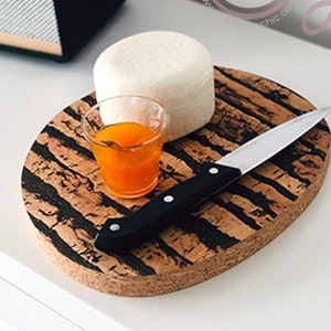 May include: A cork serving board with a black striped design, holding a small glass of orange liquid, a white cheese wheel, and a knife. The board is on a white surface. A black speaker is in the background.