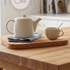 Puede incluir: Una tetera color crema, una taza de té con una cuchara y un cuenco de madera pequeño están sobre una tabla de corcho. Una pila de platos y una servilleta de lino están a la derecha. Los artículos están sobre una mesa de madera en una cocina.