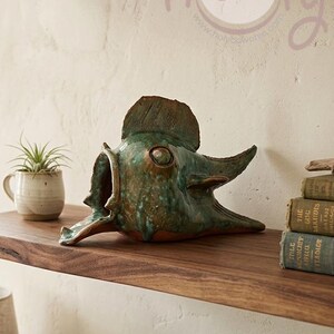 May include: A ceramic fish sculpture with a distressed green and brown finish rests on a wooden shelf. The sculpture has an open mouth and a fin on top. A small potted plant and books are also on the shelf.