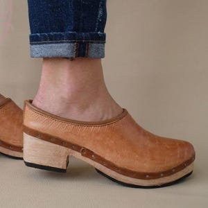 Può includere: Zoccoli in pelle marrone chiaro con tacchi in legno. Gli zoccoli hanno la punta arrotondata e sono fissati con piccole borchie metalliche. I tacchi sono alti circa 5 cm. Indossati con jeans blu.