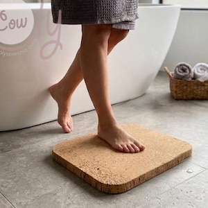 May include: A square cork bath mat on a grey tiled floor. A person stands on the mat near a white bathtub. A small woven basket with rolled towels and a potted plant are in the background.