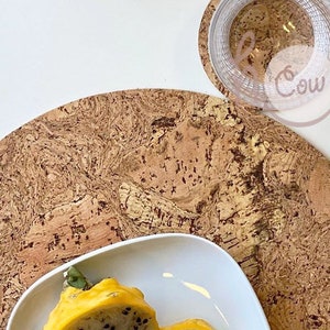 May include: A round cork trivet with a marbled pattern in shades of brown and beige. A white ceramic plate with two yellow fruits is on top of the trivet.