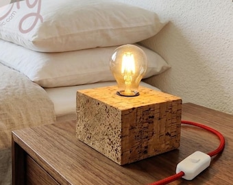 May include: A cork lamp with a lit bulb sits on a wooden bedside table. The lamp has a red cord and a white switch. The background includes a bed with white pillows and a framed print. The lamp is approximately 15 cm in height.