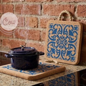 May include: A navy blue cast iron pot rests on a cork trivet with a blue floral pattern. A matching cork trivet with a rope handle stands upright. The background features a brick wall and a stovetop.