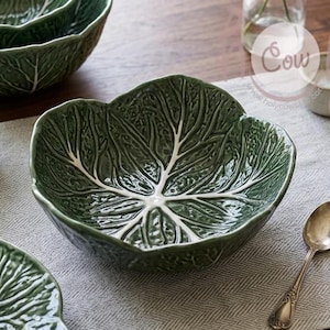 Op de afbeelding: Een verzameling groene keramische kommen en borden, gevormd en getextureerd om op koolbladeren te lijken. De set bevat kommen en borden in verschillende maten, perfect voor het serveren van salades of bijgerechten. Een lepel en een servetring zijn ook zichtbaar.