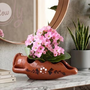 Puede incluir: Una jardinera de cerámica color terracota con forma de barco, llena de vibrantes flores rosas. La jardinera tiene un patrón de puntos blancos y diseños florales negros. El fondo incluye un espejo redondo y una planta en maceta.