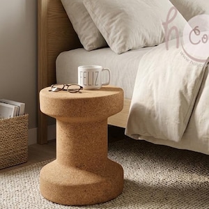 May include: A cylindrical cork side table with a spool-like shape, featuring a flat top surface. Resting on the table is a white ceramic mug with the word FELIZ printed in black lettering and a pair of dark-rimmed eyeglasses.