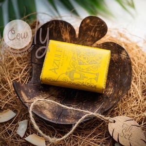 Peut inclure: Un savon jaune avec l'inscription "AZULEK" dans un plat en noix de coco marron, posé sur un lit de paille. La présentation comprend des copeaux de noix de coco et une étiquette cadeau en bois.