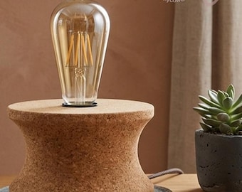 May include: A table lamp with a clear Edison-style bulb on a cork base. The lamp sits on a wooden surface, with a small succulent plant in a dark grey pot to the right. The lamp's base is approximately 15 cm in diameter.