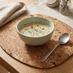 May include: A light green bowl filled with creamy soup, garnished with herbs and pepper, sits on a cork placemat. A silver spoon rests beside the bowl. A bottle and a small jar with rosemary sprigs are in the background.