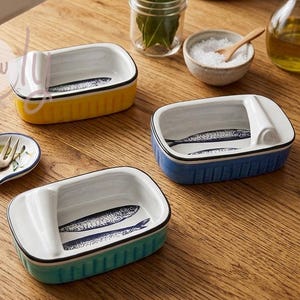 May include: Three ceramic sardine dishes in yellow, blue, and teal, each with a white interior and a sardine illustration. A small dish with a fork and a striped cloth are also present on a wooden table.