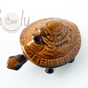 Puede incluir: Una figurita de tortuga de cerámica marrón con una tortuga más pequeña en su espalda. La tortuga tiene un caparazón detallado con un patrón texturizado.