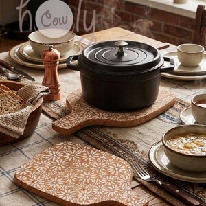 Puede incluir: Una olla negra de hierro fundido con tapa sobre un salvamanteles de madera decorativo con un estampado floral blanco. La mesa está puesta con cuencos, platos y un molinillo de pimienta, lo que sugiere una comida.