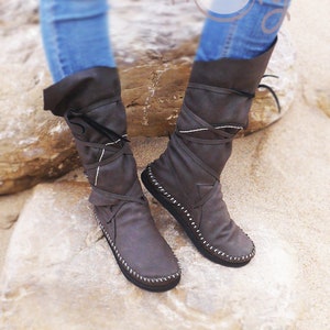 May include: A pair of brown leather boots with a unique design. The boots have a folded-over top, wrap-around laces, and white stitching details. The boots are worn with blue jeans.