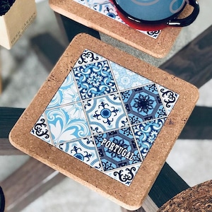 Pode incluir: Duas bases de cortiça quadradas com um design de azulejo português em tons de azul e branco. Uma caneca azul e vermelha está em cima de uma das bases. As bases estão numa mesa de vidro.