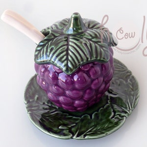 May include: A ceramic honey pot shaped like a purple blackberry with a green leaf lid and a wooden spoon. The pot sits on a matching green leaf plate.