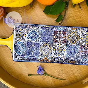 Puede incluir: Una bandeja rectangular para servir con un asa amarilla y un diseño estampado en azul y blanco. El patrón recuerda a los azulejos tradicionales portugueses. La bandeja está sobre una superficie de madera.