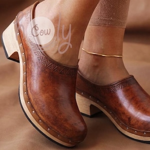 Puede incluir: Zuecos de cuero marrón con tacón y suela de madera. Los zuecos tienen un patrón decorativo en la parte superior y están asegurados con pequeños tachuelas metálicas alrededor de la suela. La imagen muestra los zuecos en los pies de una persona.