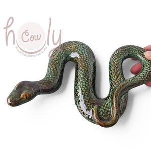 Può includere: Una statuetta di serpente in ceramica lucida caratterizzata da un motivo a squame sovrapposte e testurizzate in tonalità verde bosco scuro, oliva e marrone. Il serpente è raffigurato in una posa sinuosa a forma di S con finitura liscia.