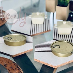 Puede incluir: Tres posavasos hexagonales de corcho con rayas blancas y negras. Cada posavasos tiene un objeto diferente en la parte superior: una lata dorada, una olla de cerámica blanca y otra lata dorada.