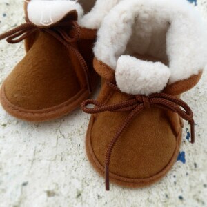 May include: A pair of brown baby booties with white fluffy lining and brown laces. The booties are made of a soft material and are designed to keep a baby's feet warm and comfortable. The style is reminiscent of moccasins.